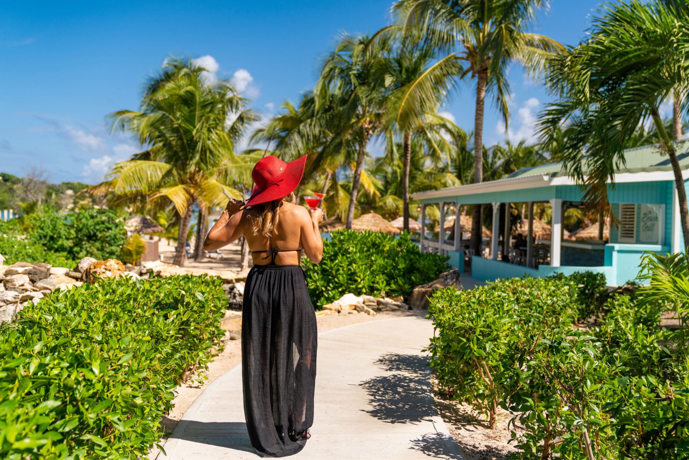 A guest in a sun hat walking toward a beachside bar through tropical gardens